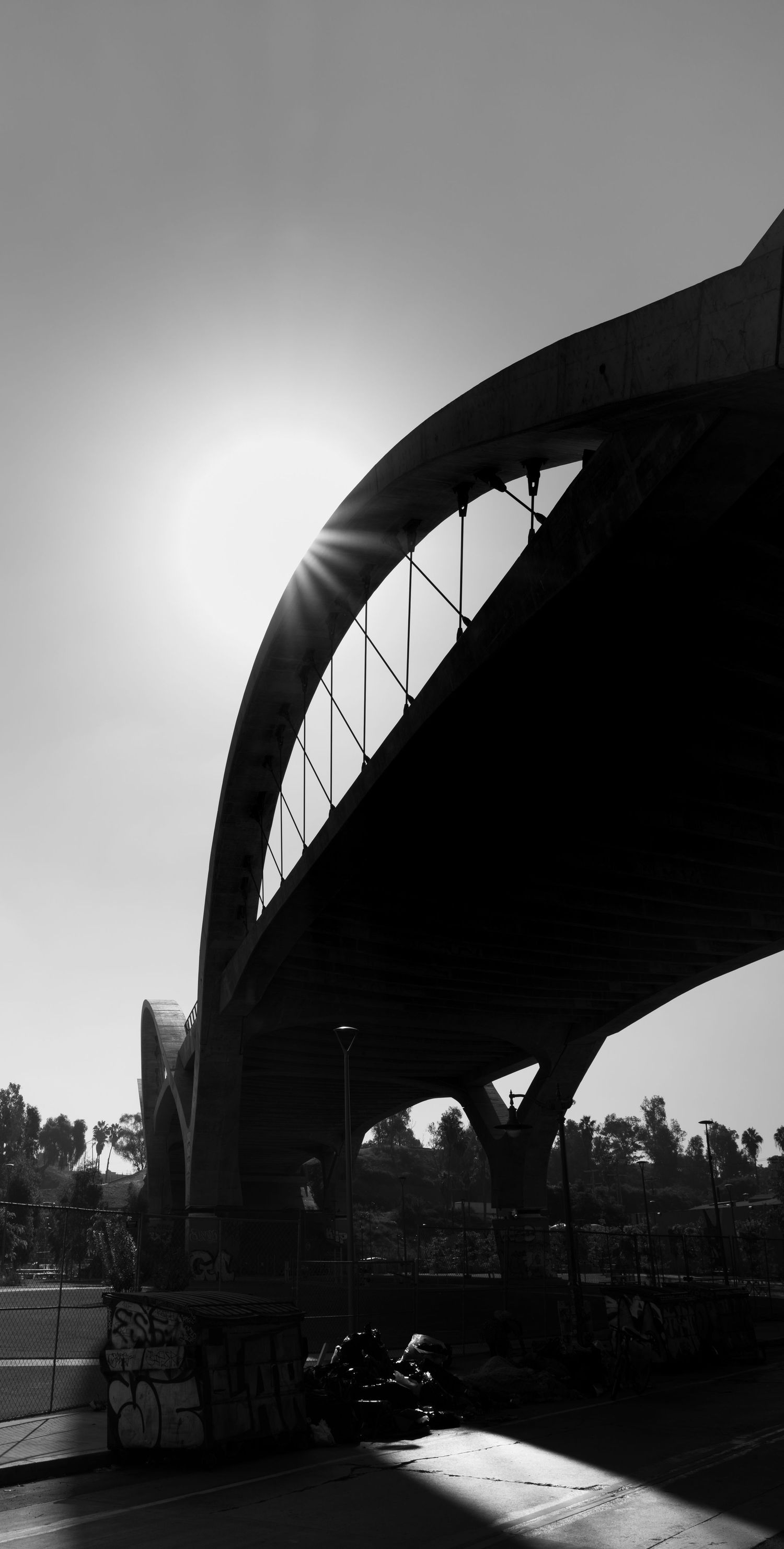 Black and white silhouette of an arch bridge against a bright sun, with graffiti-covered walls and trees in the background.
