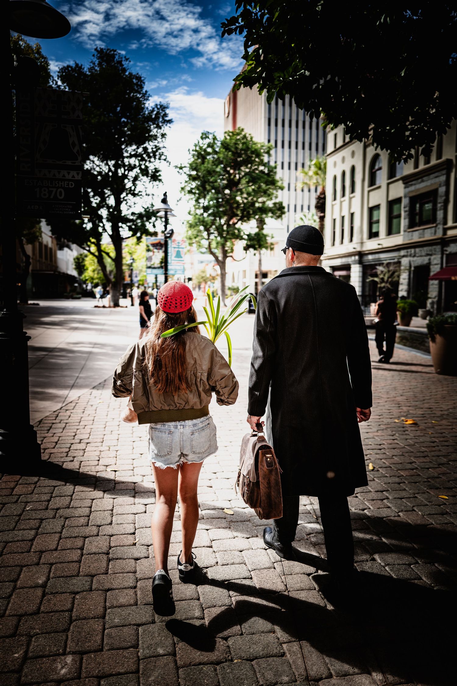 A girl in a red cap and denim shorts walks beside a man in a long dark coat along a sunlit brick sidewalk in a tree-lined city street.