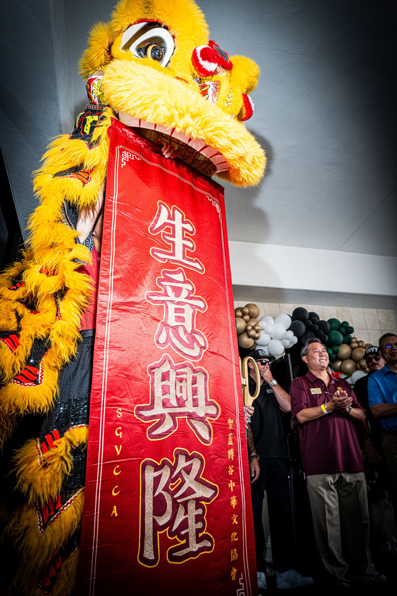 A yellow lion dance costume holds a red banner with gold Chinese characters while a smiling man applauds at a festive grand opening event.
