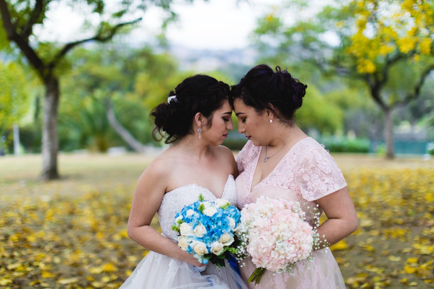 Two brides in white and pink gowns hold floral bouquets and touch foreheads in a romantic outdoor park setting.