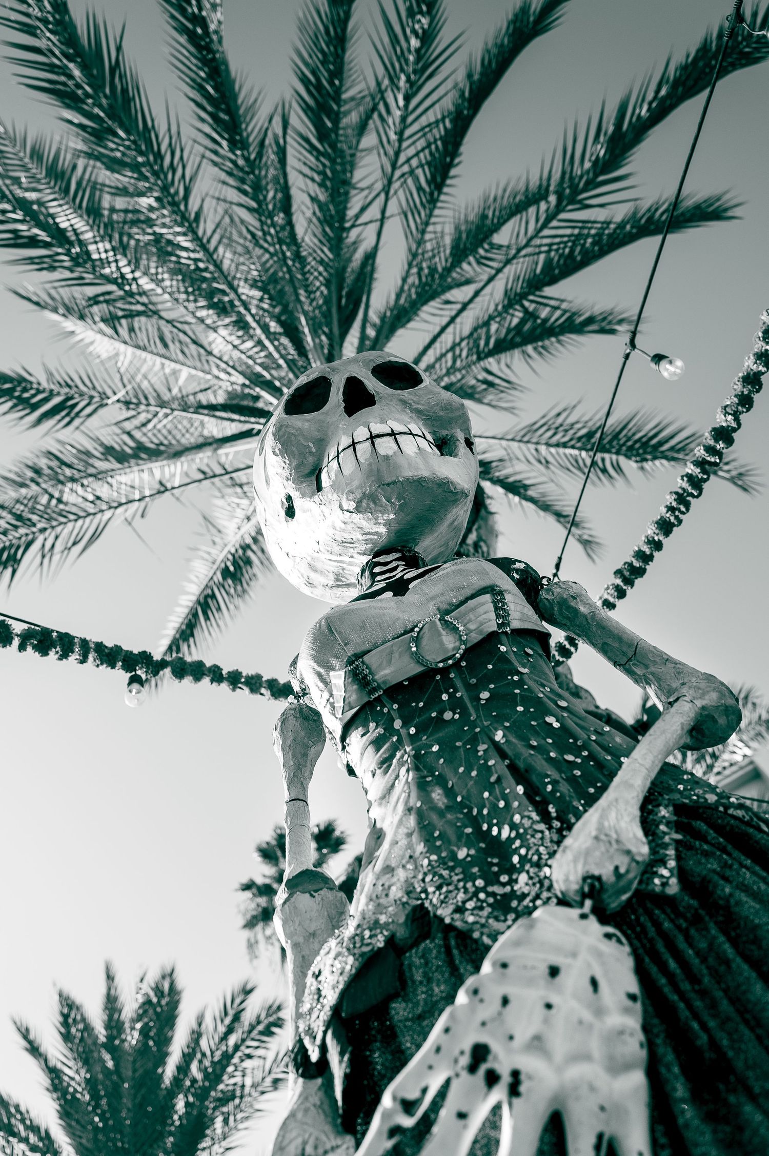 Low-angle black and white photo of a skeleton statue under a palm tree at a festival.