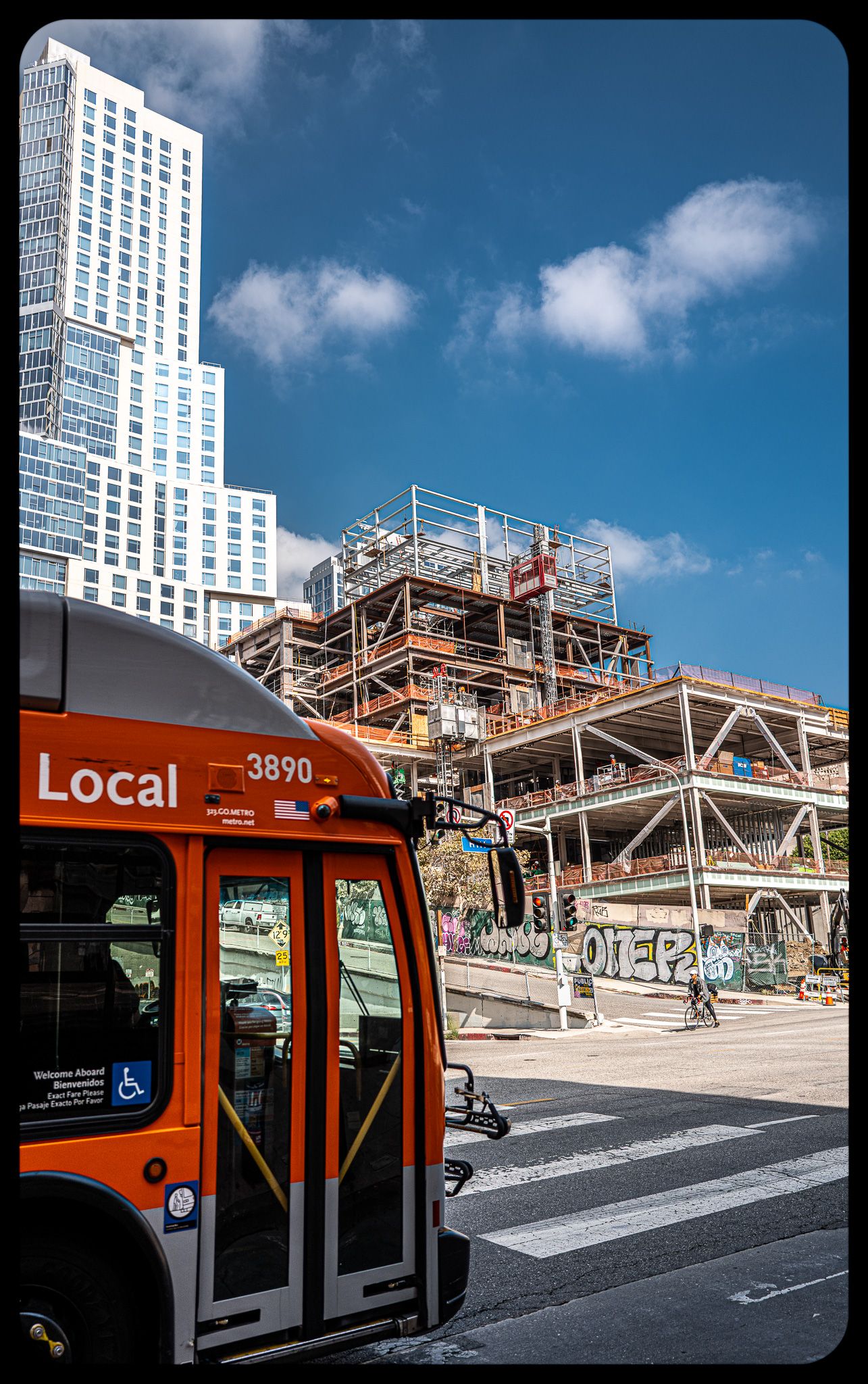 Orange local city bus number 3890 at intersection near active construction site and skyscrapers.