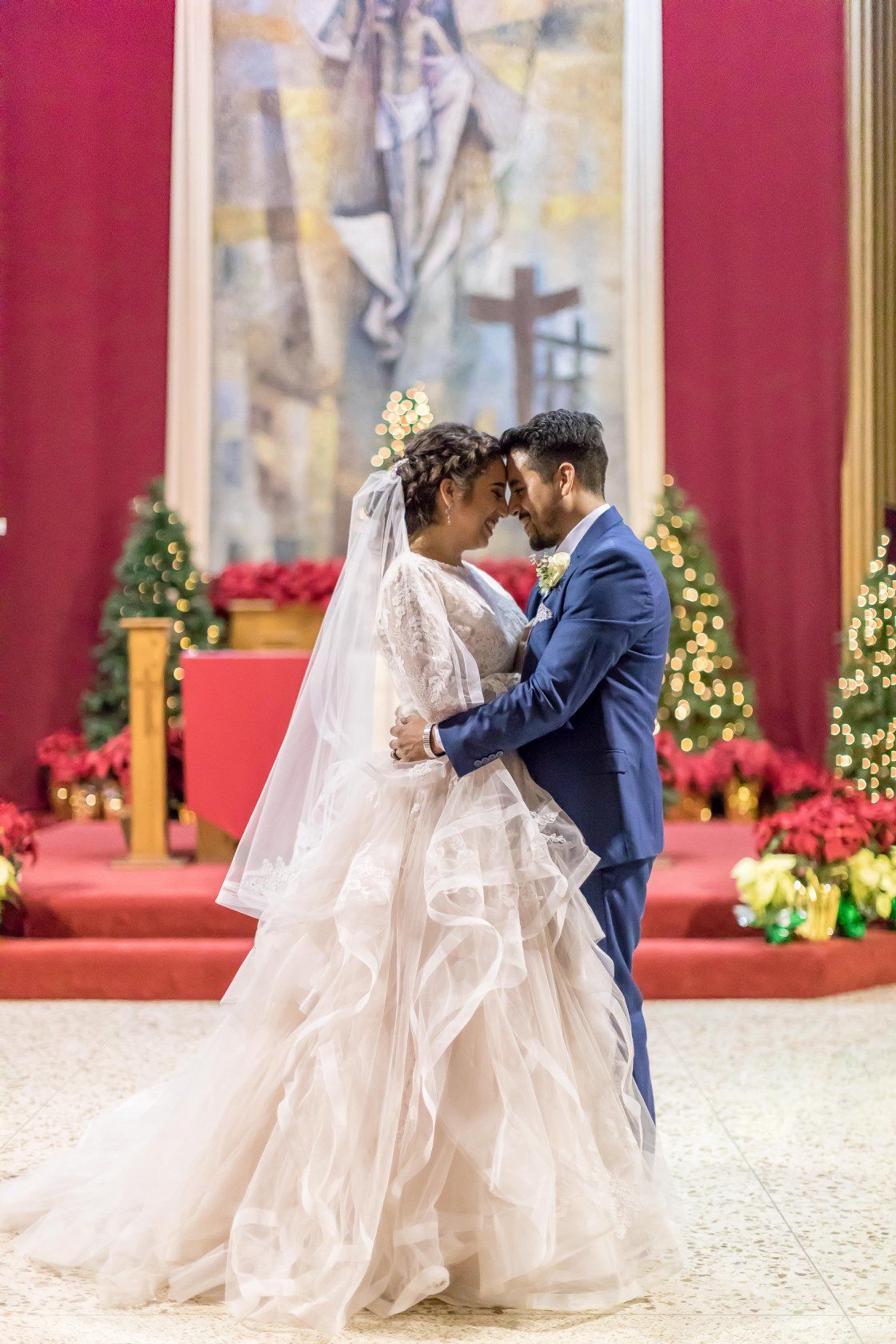 Bride and groom share romantic forehead touch in church decorated with Christmas trees and poinsettias.