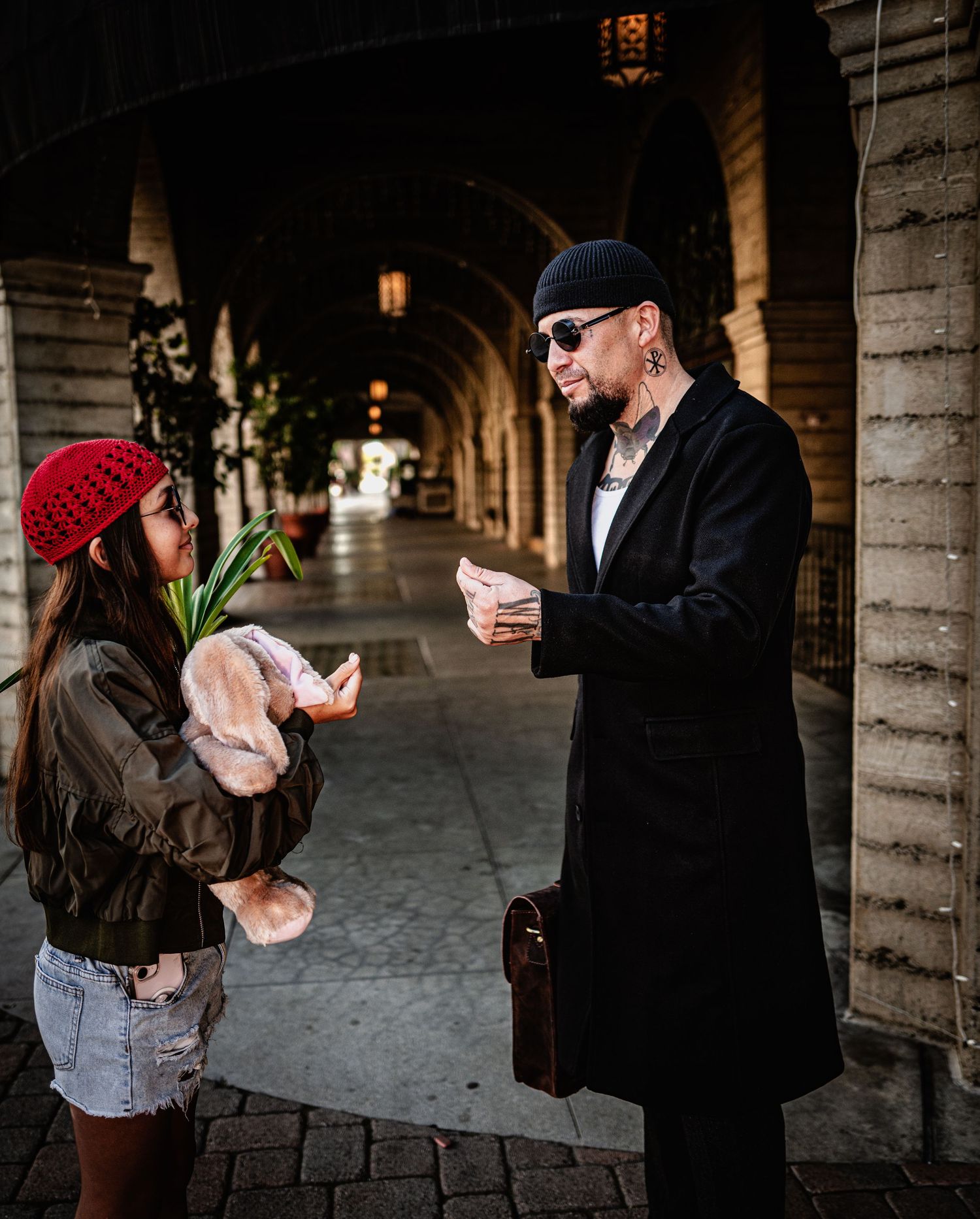 A man in a black coat, beanie and sunglasses talks with a woman in a red knit hat holding a plush toy under a stone arcade.