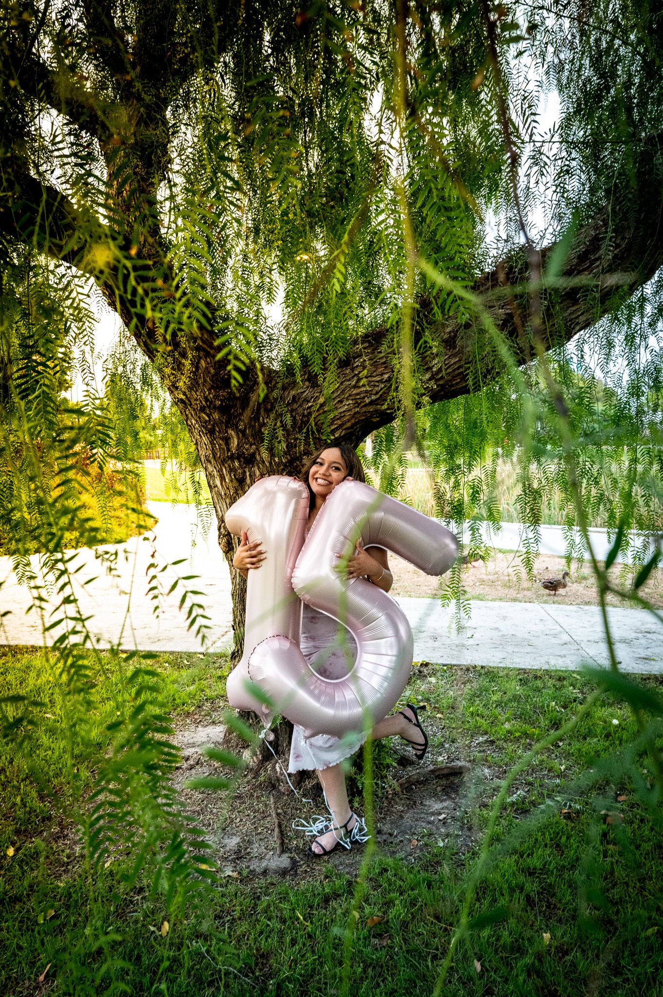 A smiling woman hugs large number 15 balloons under a weeping willow tree outdoors.