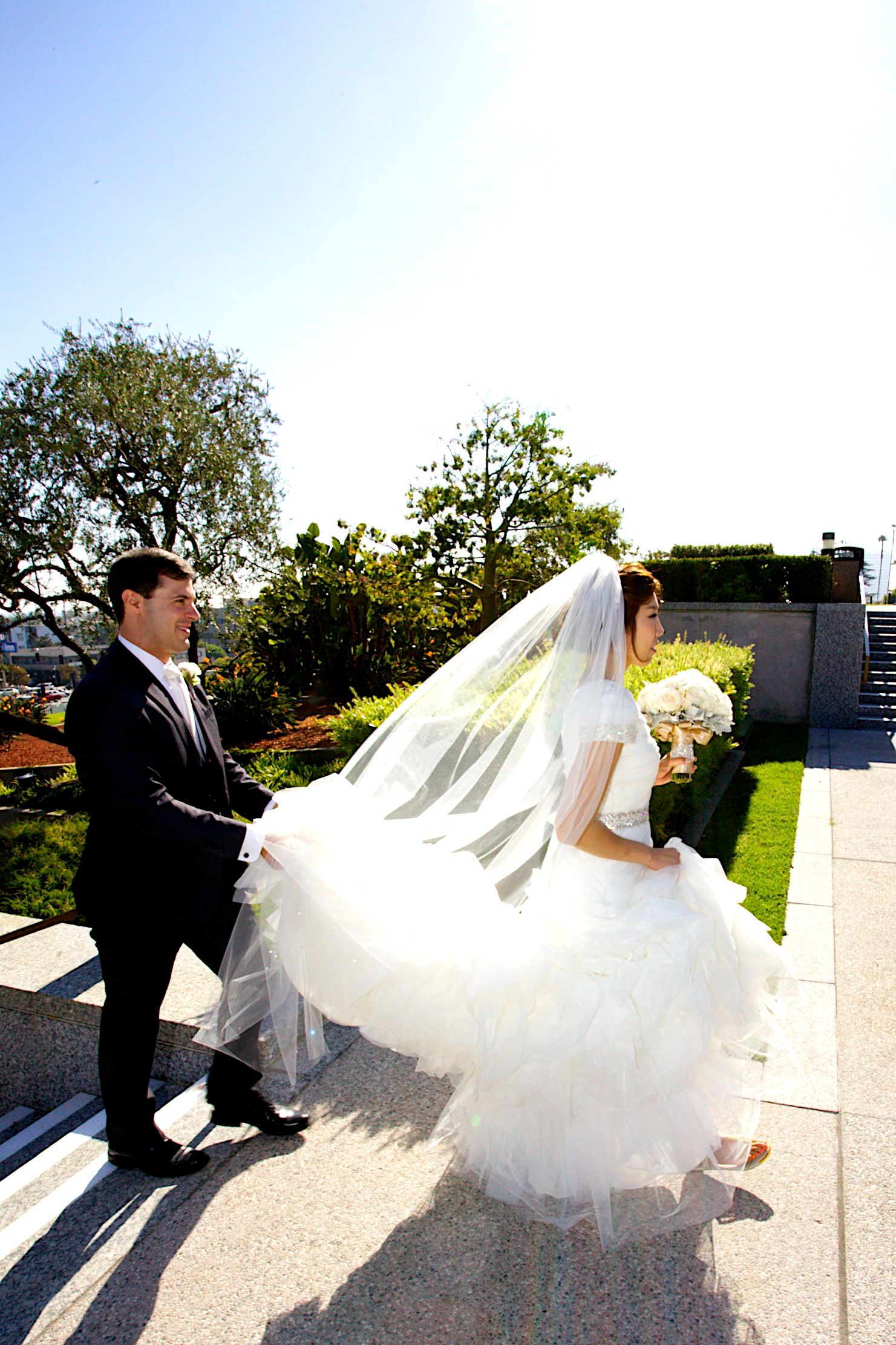 Bride in white gown and veil walks with groom in black tuxedo on sunny outdoor terrace surrounded by lush greenery.