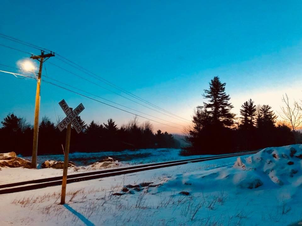Snowy railroad crossing at dusk with illuminated signal, power lines, and silhouetted evergreen trees under a blue sky.