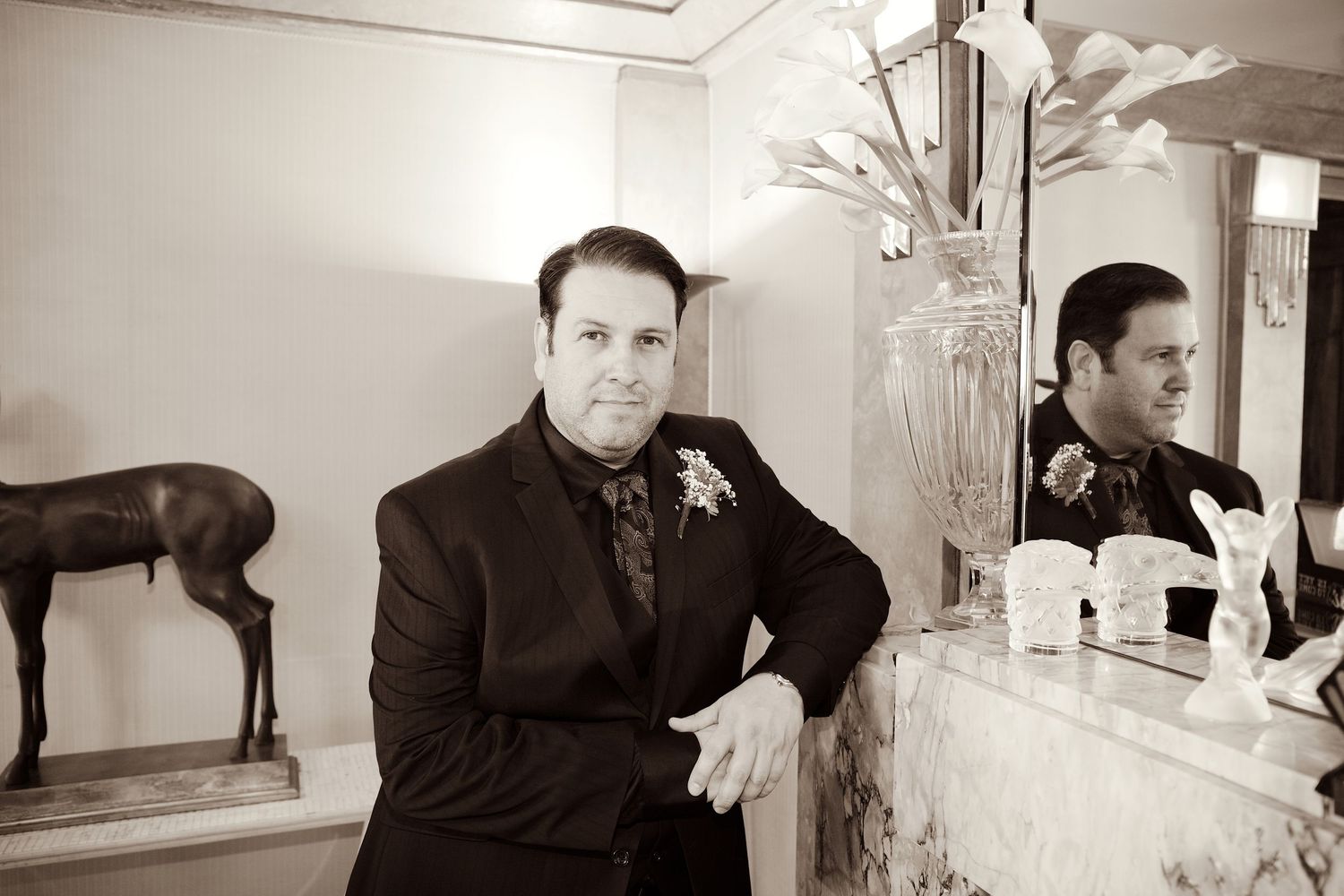 Elegant man in black suit with boutonniere leaning on a marble mantle beside a horse sculpture.