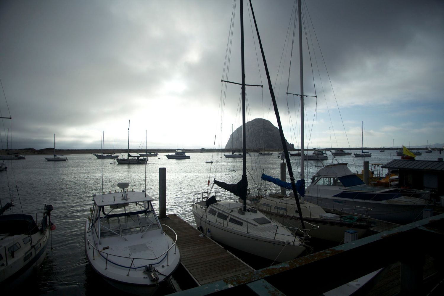 Sailboats docked at a marina with Morro Rock silhouetted against a dramatic cloudy sky at dusk.