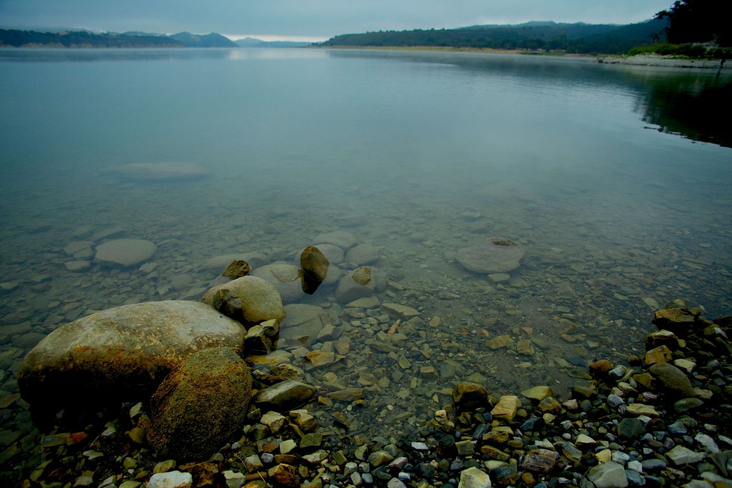 Clear shallow lake with smooth rocks and pebbles on shoreline, misty mountains in background on a calm overcast day.