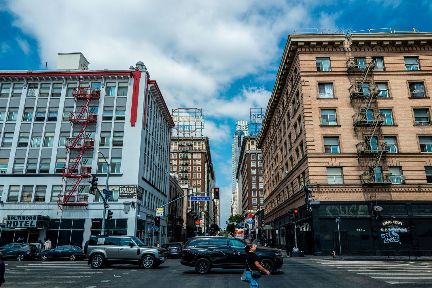 Busy urban street intersection with tall buildings, fire escapes, and cars in a city downtown area under a partly cloudy sky.