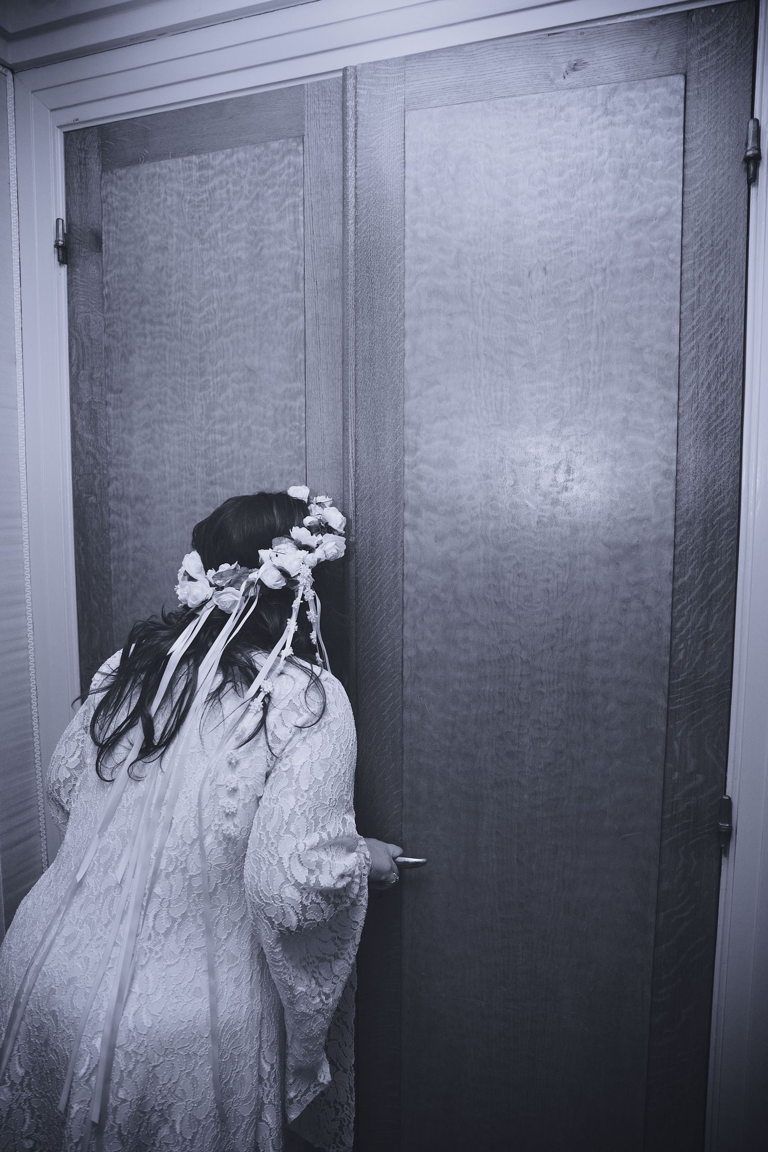 Black and white photo of a bride in lace dress and floral crown standing at a door.