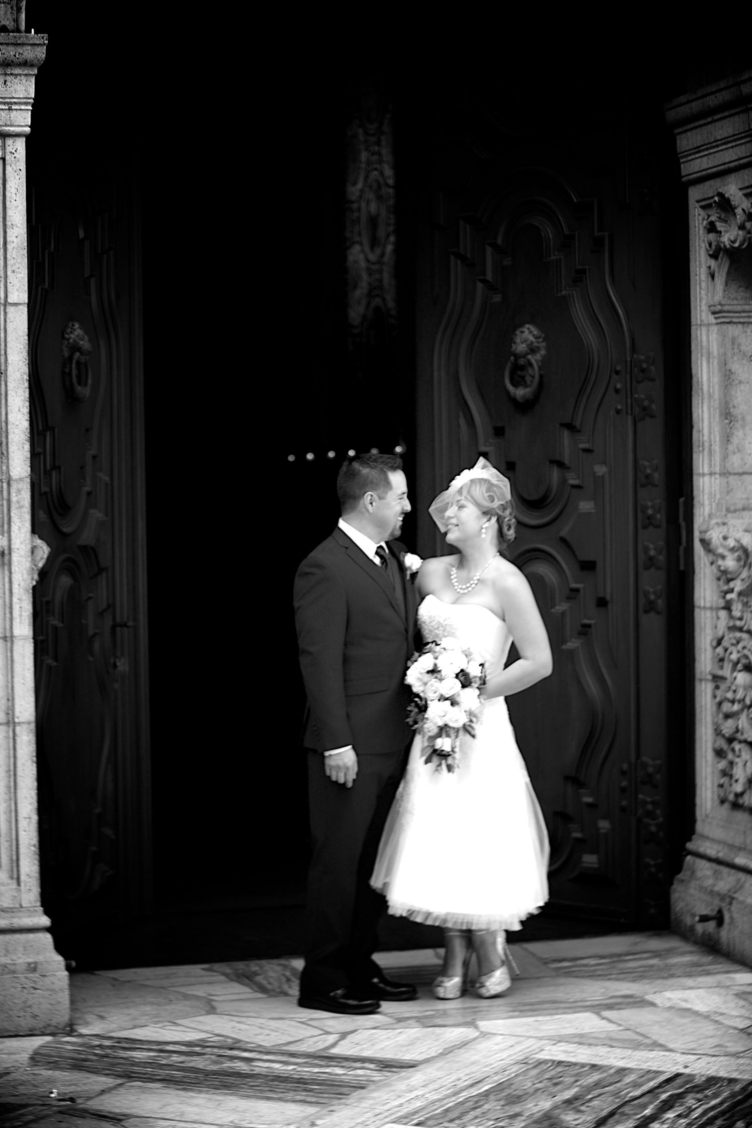 Black and white wedding photo of bride and groom standing before ornate church doors, bride holding bouquet.