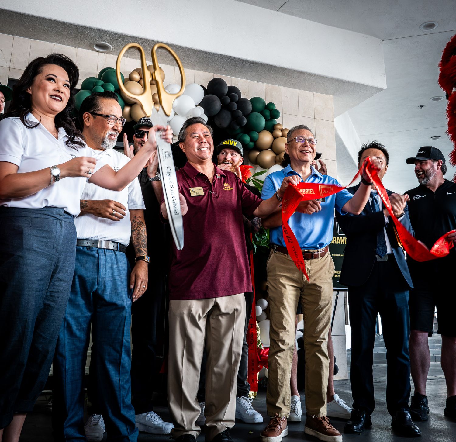 Smiling group holds oversized golden scissors to cut a red ribbon at a grand opening ceremony with green, black and gold balloons.