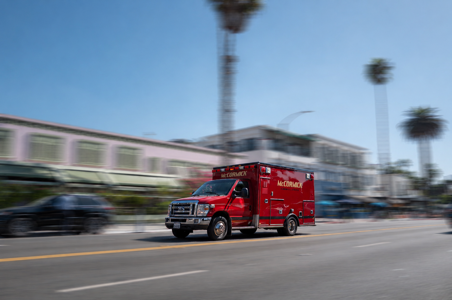 Red ambulance speeding down urban street with motion blur, palm trees and buildings in background.