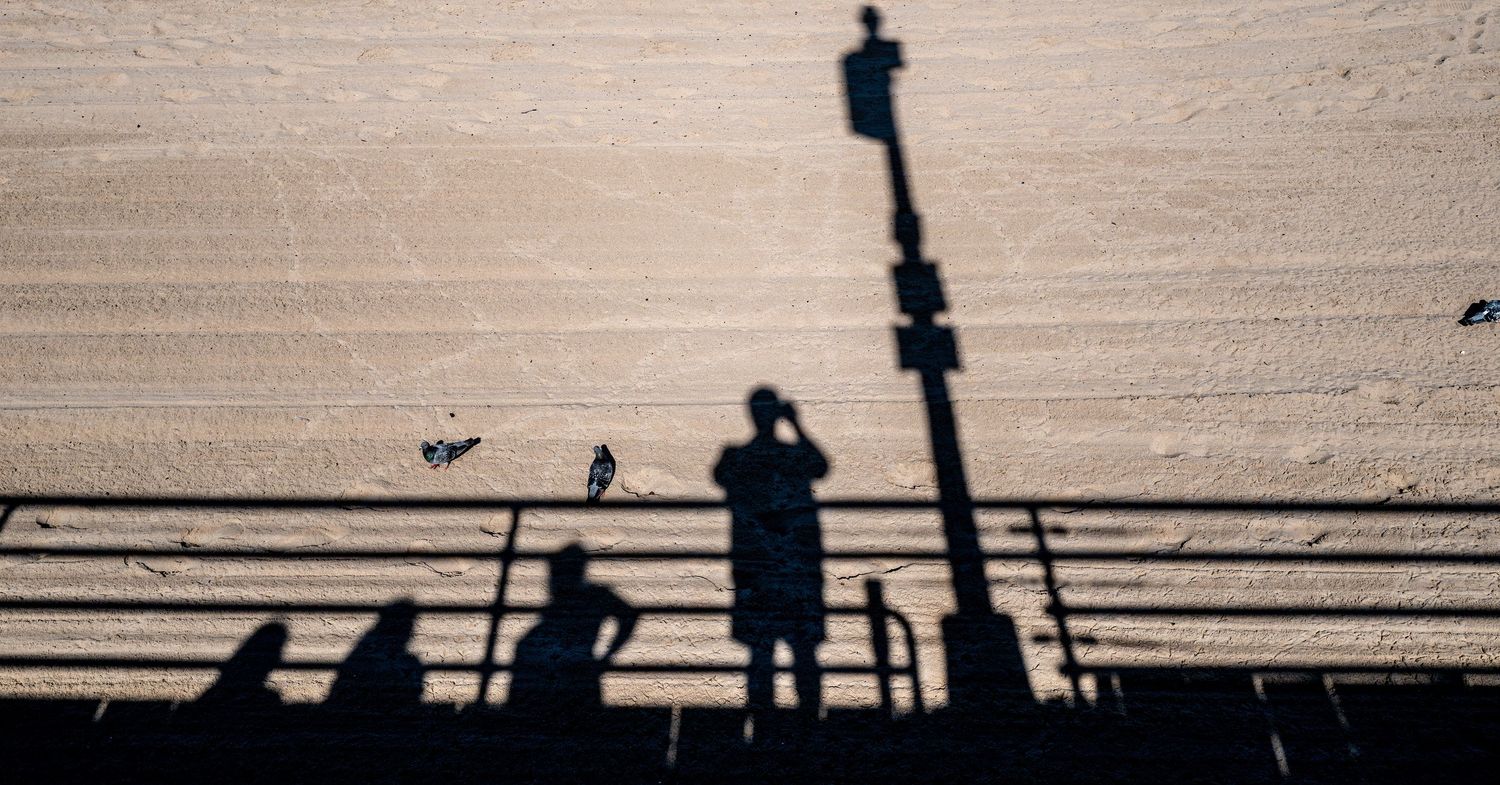 Shadows of people on a boardwalk cast onto a sandy beach in bright sunlight.