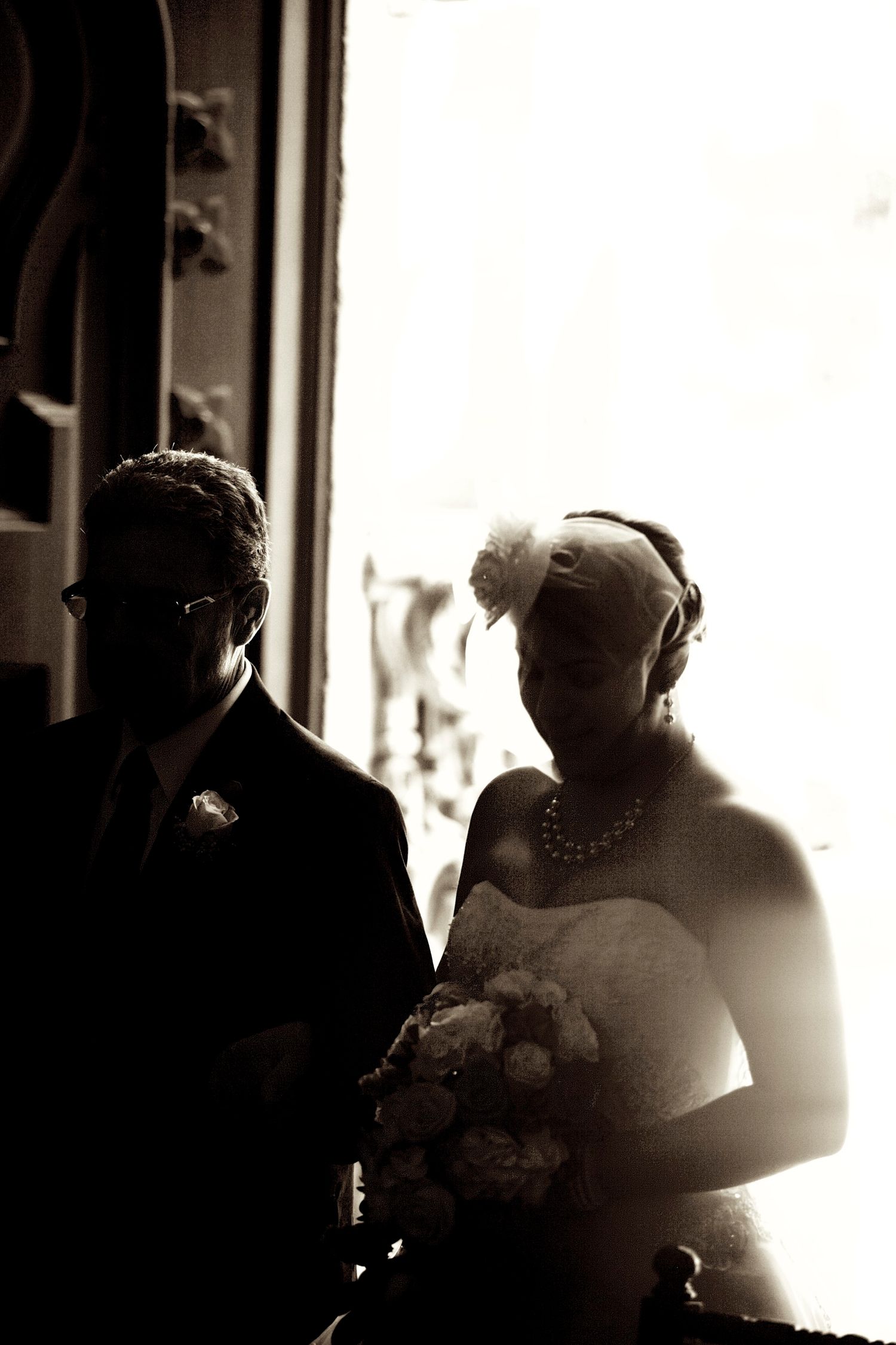 Sepia-toned wedding photo of bride in pearl necklace holding bouquet beside groom in suit, dramatic backlit silhouette.