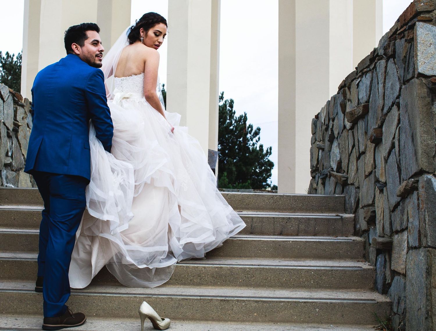 Groom helps bride carry her white gown up stone steps at elegant outdoor wedding venue.