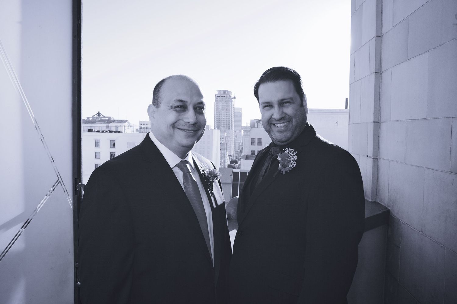 Two men in formal suits smile together on urban rooftop balcony with city skyline in black and white.