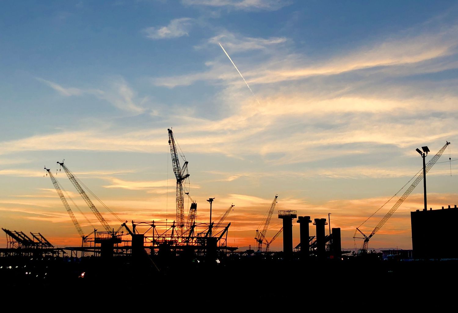 Silhouette of multiple construction cranes against a vibrant orange and blue sunset sky.