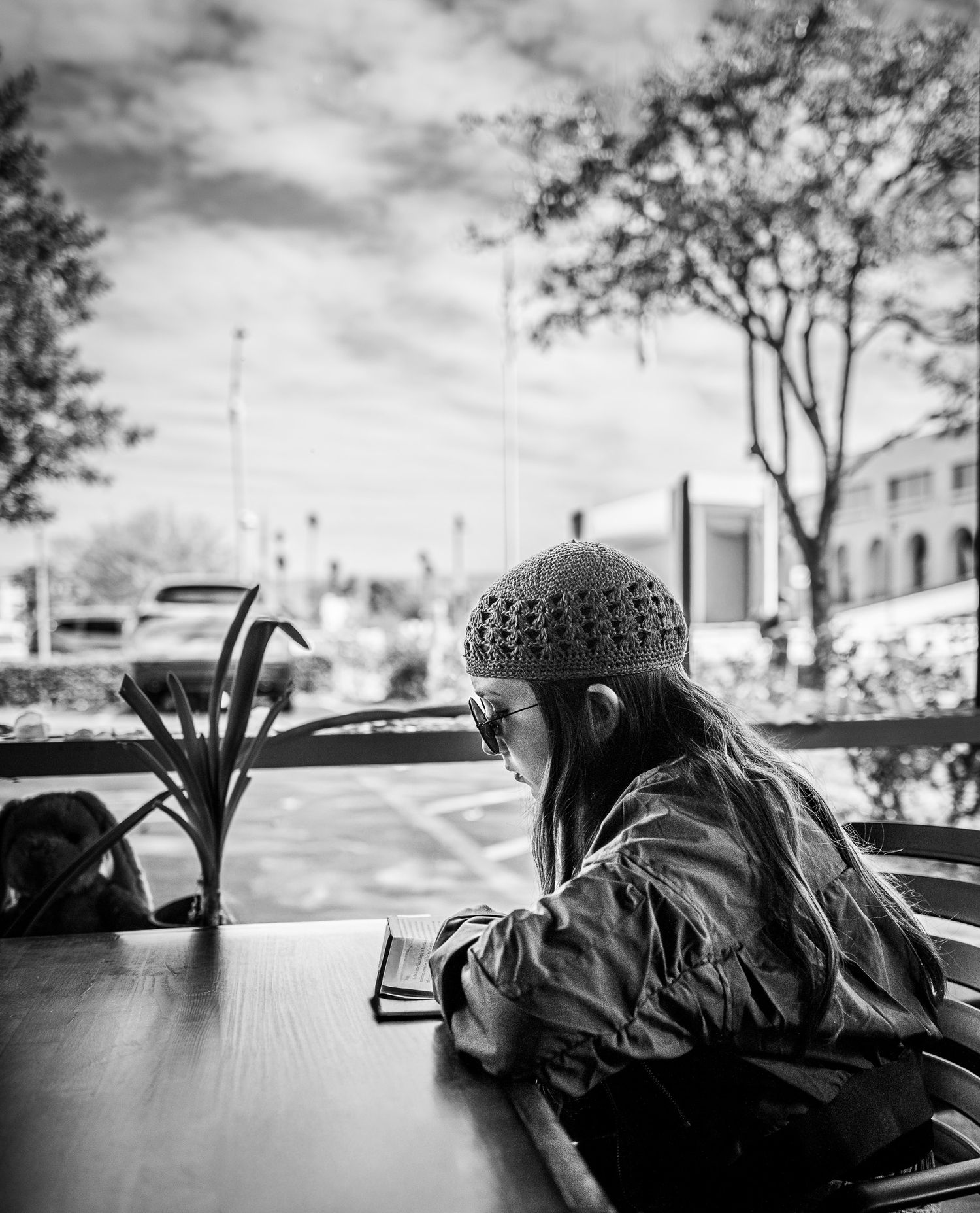 Black and white photo of a girl in a knit beanie and sunglasses sitting at an outdoor cafe table, looking down at a phone.