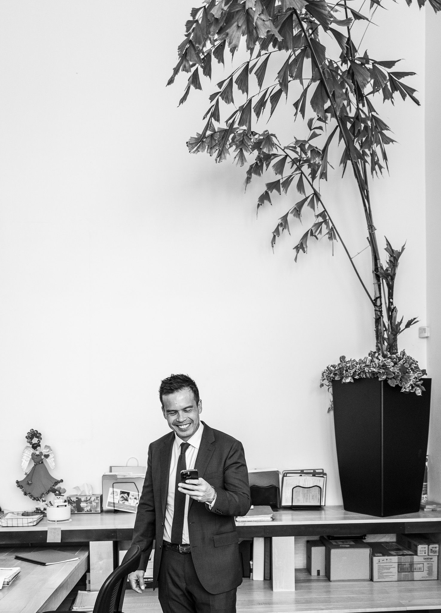 Smiling man in a suit and tie looks at his smartphone beside a desk and a tall potted plant in a bright black-and-white office.