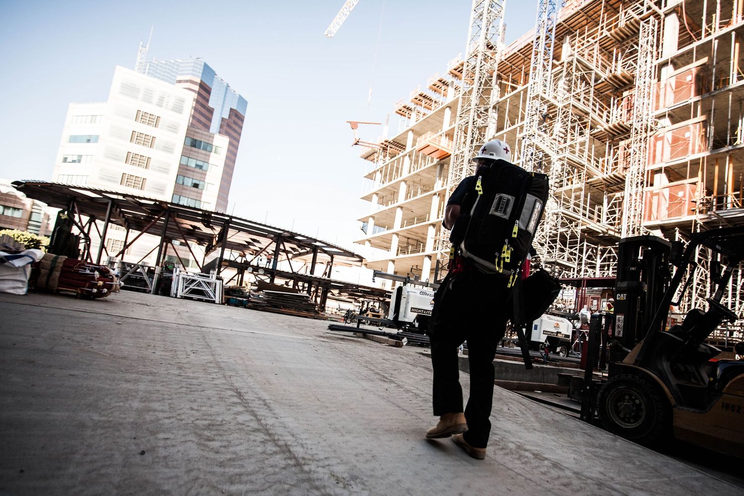 Construction worker walking toward large multi-story building frame with forklift at active job site.