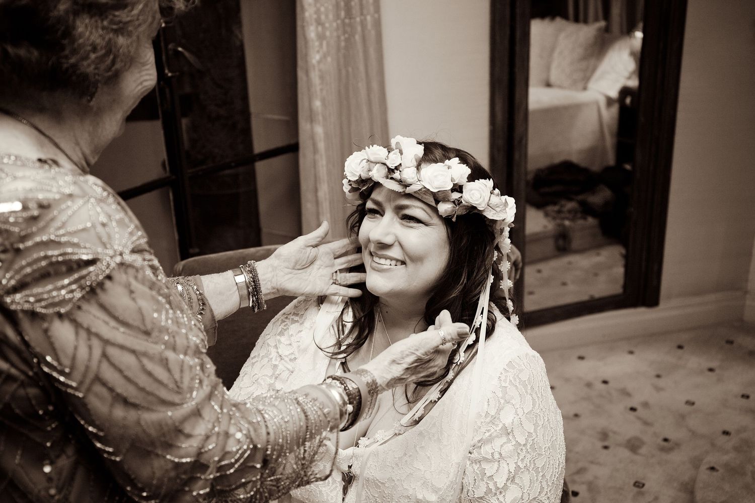 A bride smiling as someone places a floral crown on her head in sepia tones.