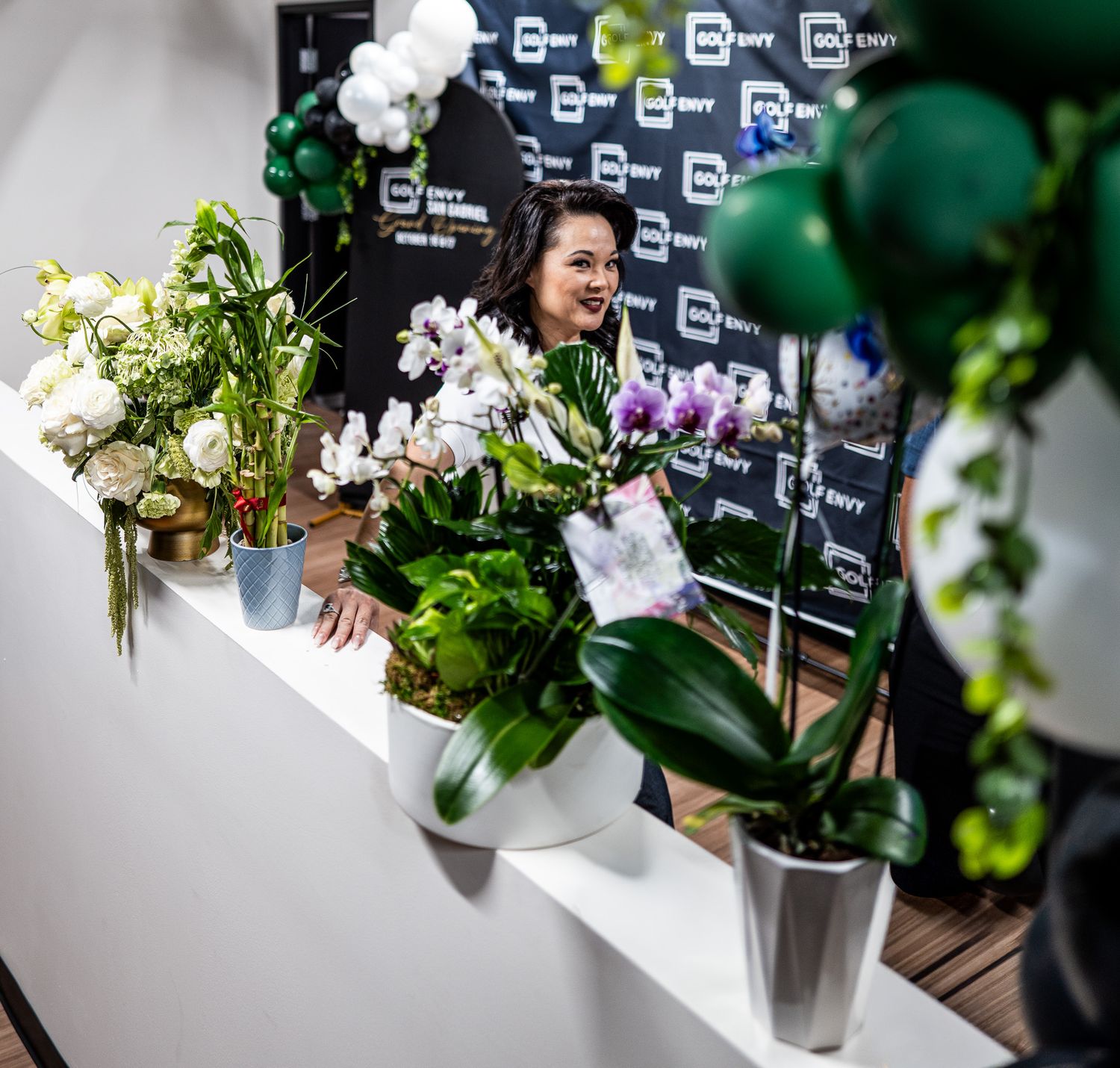 Woman smiling behind a white counter filled with orchids and green plants, with green balloons and a branded backdrop behind her.