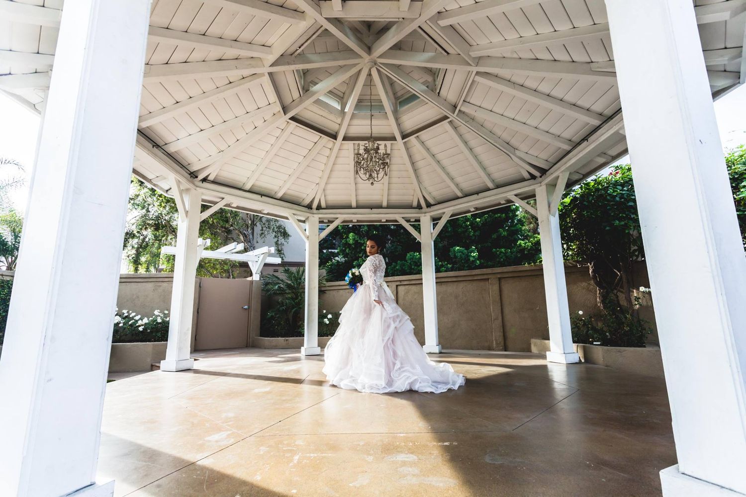 Bride in a flowing white gown stands under an elegant white gazebo with chandelier, surrounded by lush greenery.