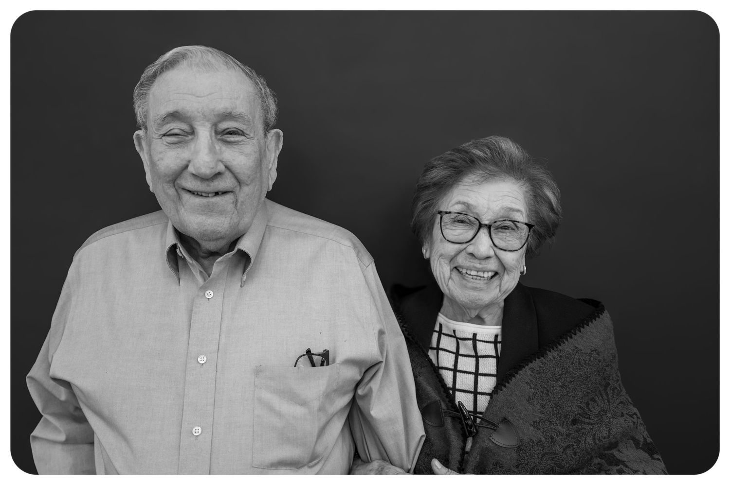 Black and white portrait of a smiling elderly couple standing together against a dark background.