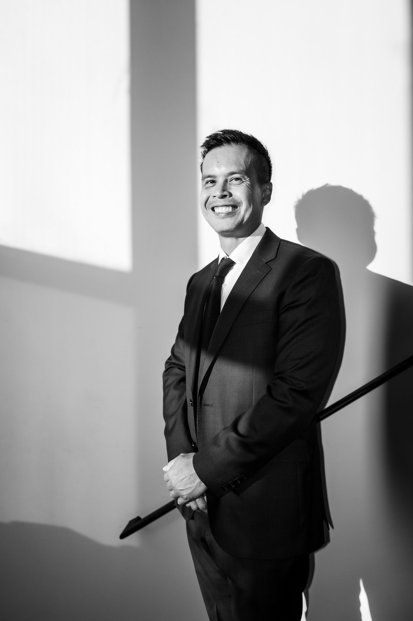 Black and white portrait of a smiling man in a dark suit and tie standing beside a railing in dramatic window light.