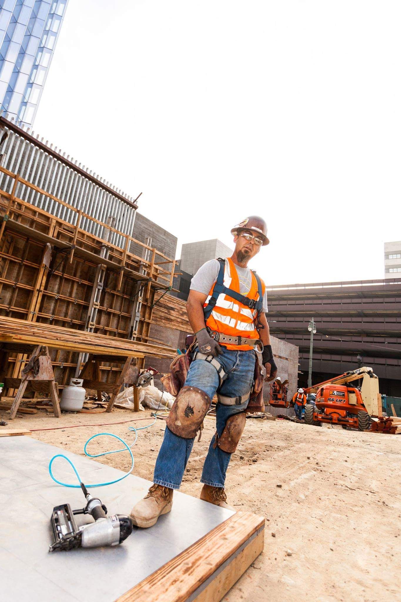 Construction worker in orange safety vest and hard hat standing on job site with nail gun on ground.