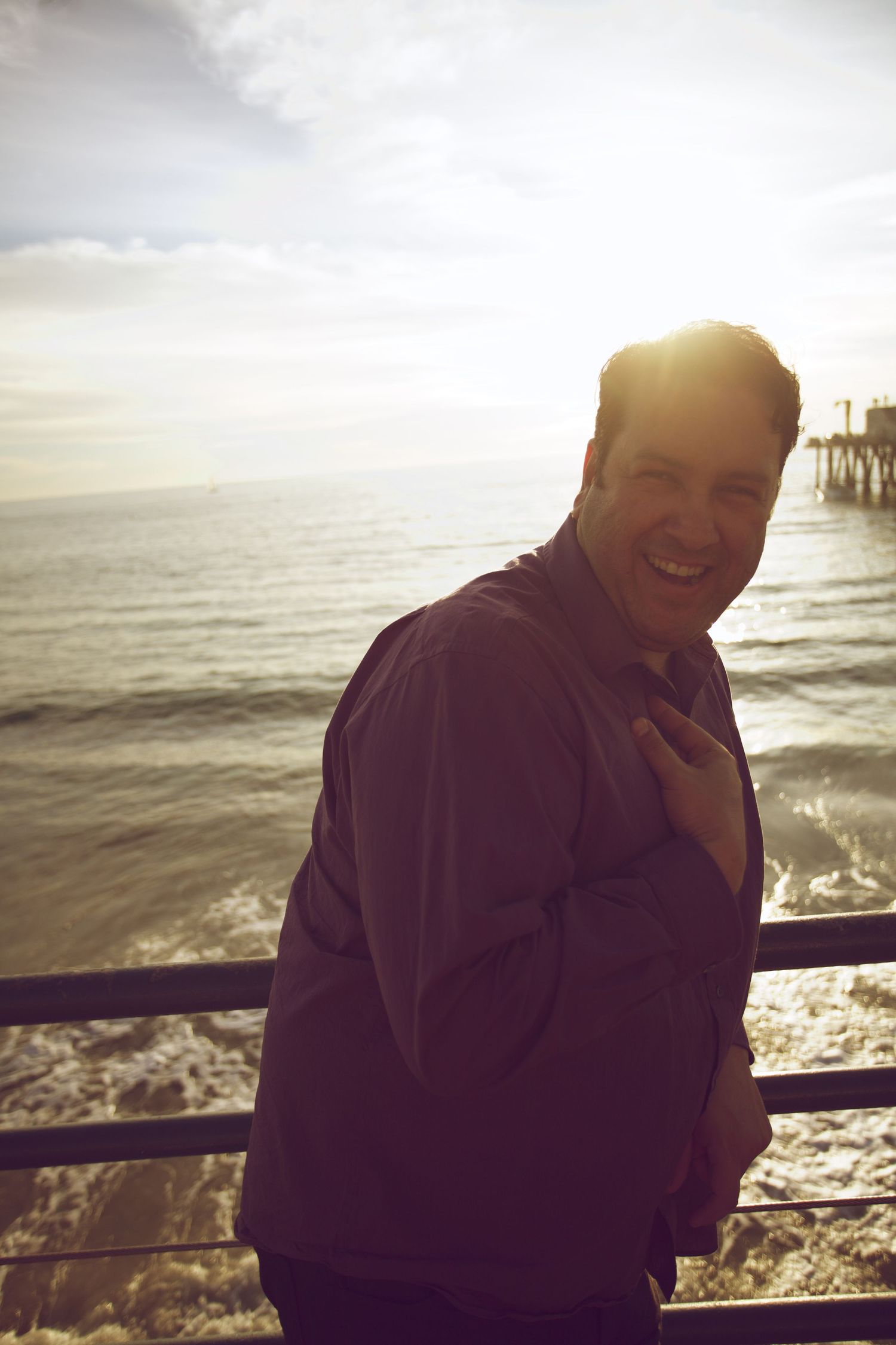 A man smiling at the ocean pier during golden hour sunset, wearing a purple jacket.
