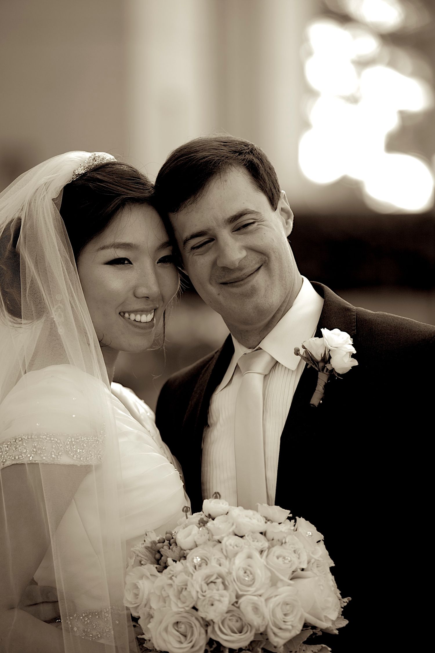 Smiling bride and groom pose together in sepia-toned wedding portrait, bride holding bouquet of white roses.
