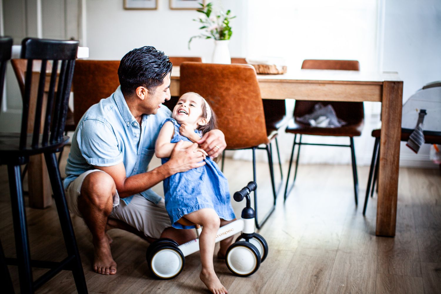 Father crouching on floor hugging laughing little girl near balance bike and dining table at home.