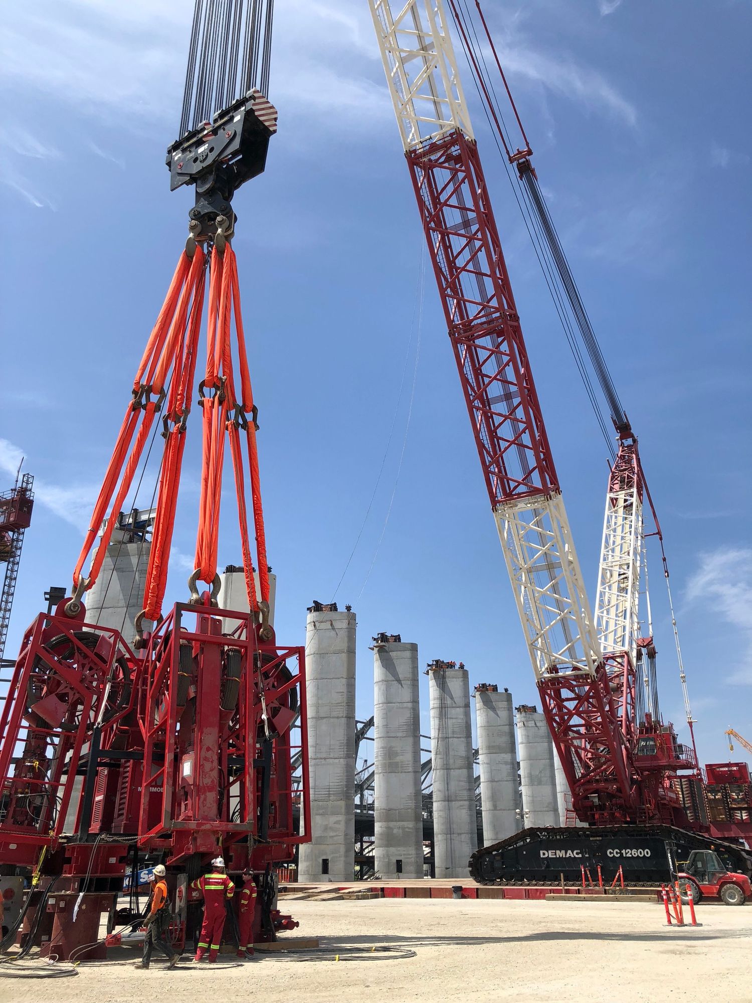 Large red crawler crane lifting heavy equipment at a construction site with concrete columns under blue sky.