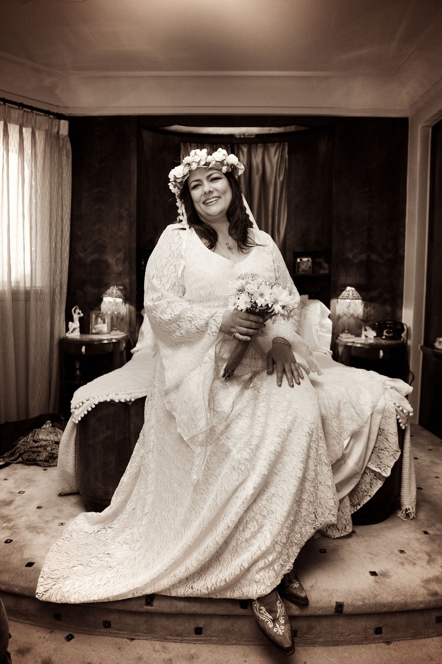 Elegant bride in vintage lace gown and floral crown sits smiling with bouquet in sepia-toned portrait.