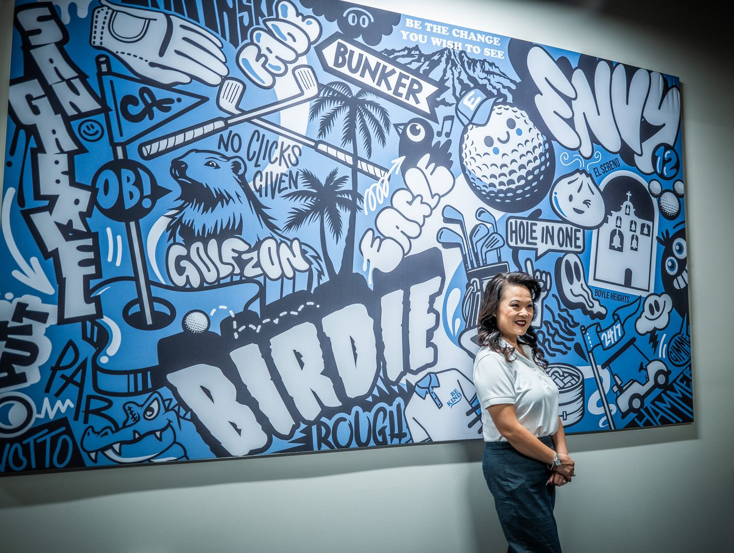 Smiling woman in a white top and jeans stands beside a blue and white golf-themed mural with the words Birdie, Bunker and Envy.
