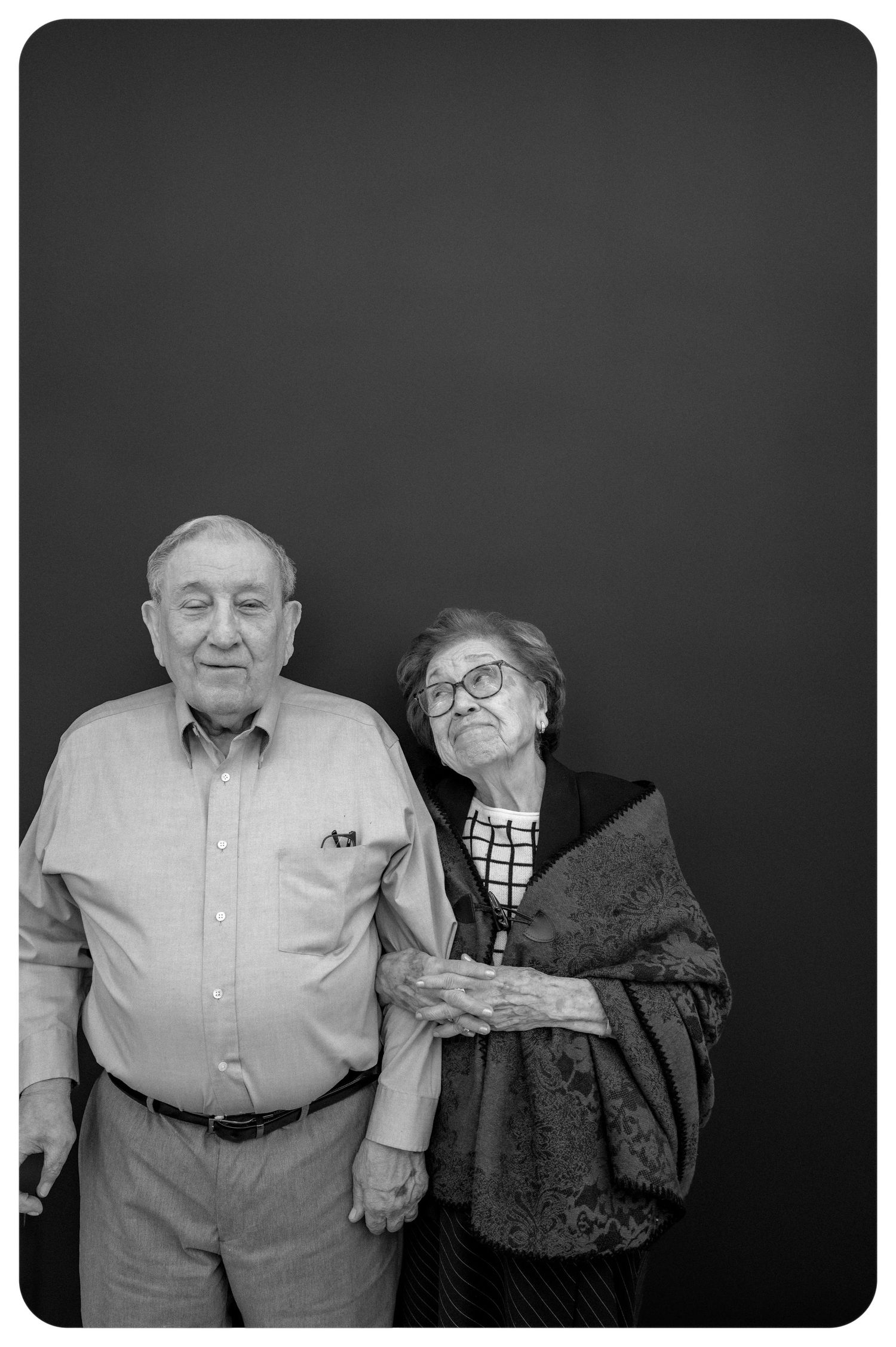 Elderly couple posing together arm in arm against a dark background in a black and white portrait.