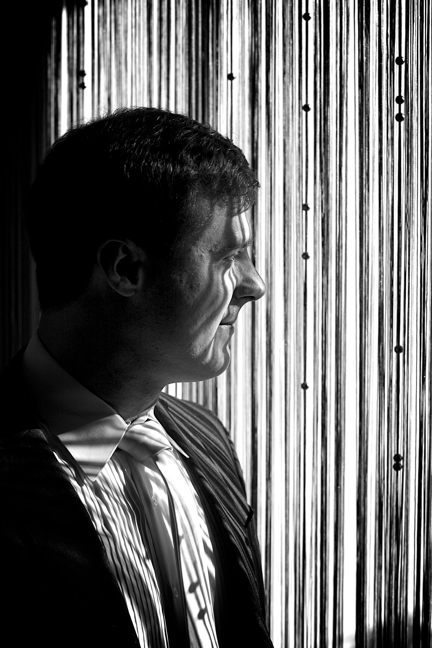 Black and white profile portrait of a man in a striped tie standing before a beaded curtain backdrop.
