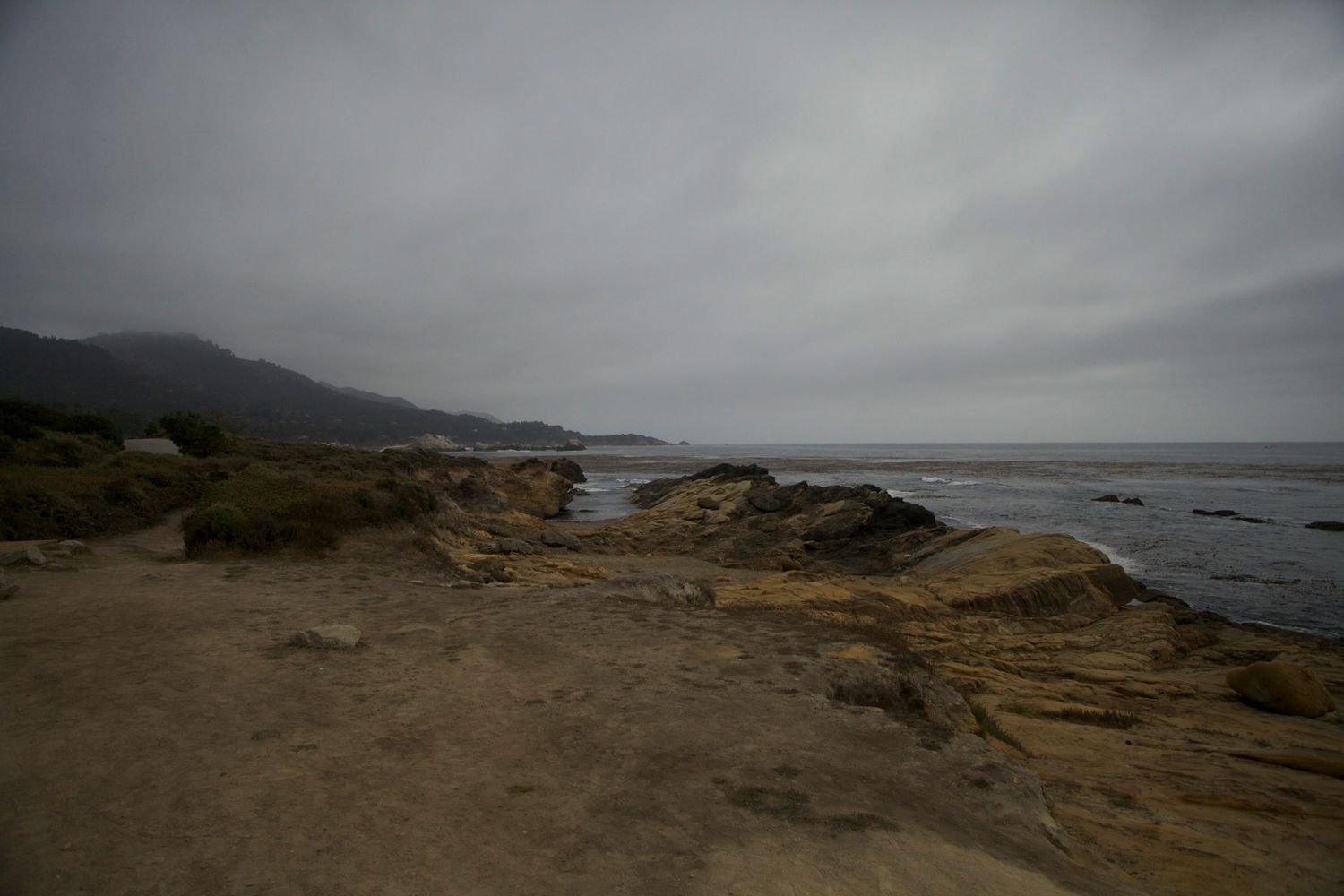 Rugged coastal rocky shoreline under overcast skies with misty mountains in the background.
