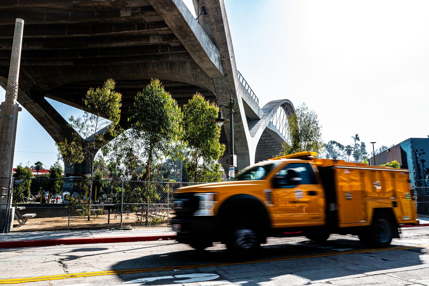 Yellow utility truck driving under a large concrete bridge overpass in an urban area on a sunny day.