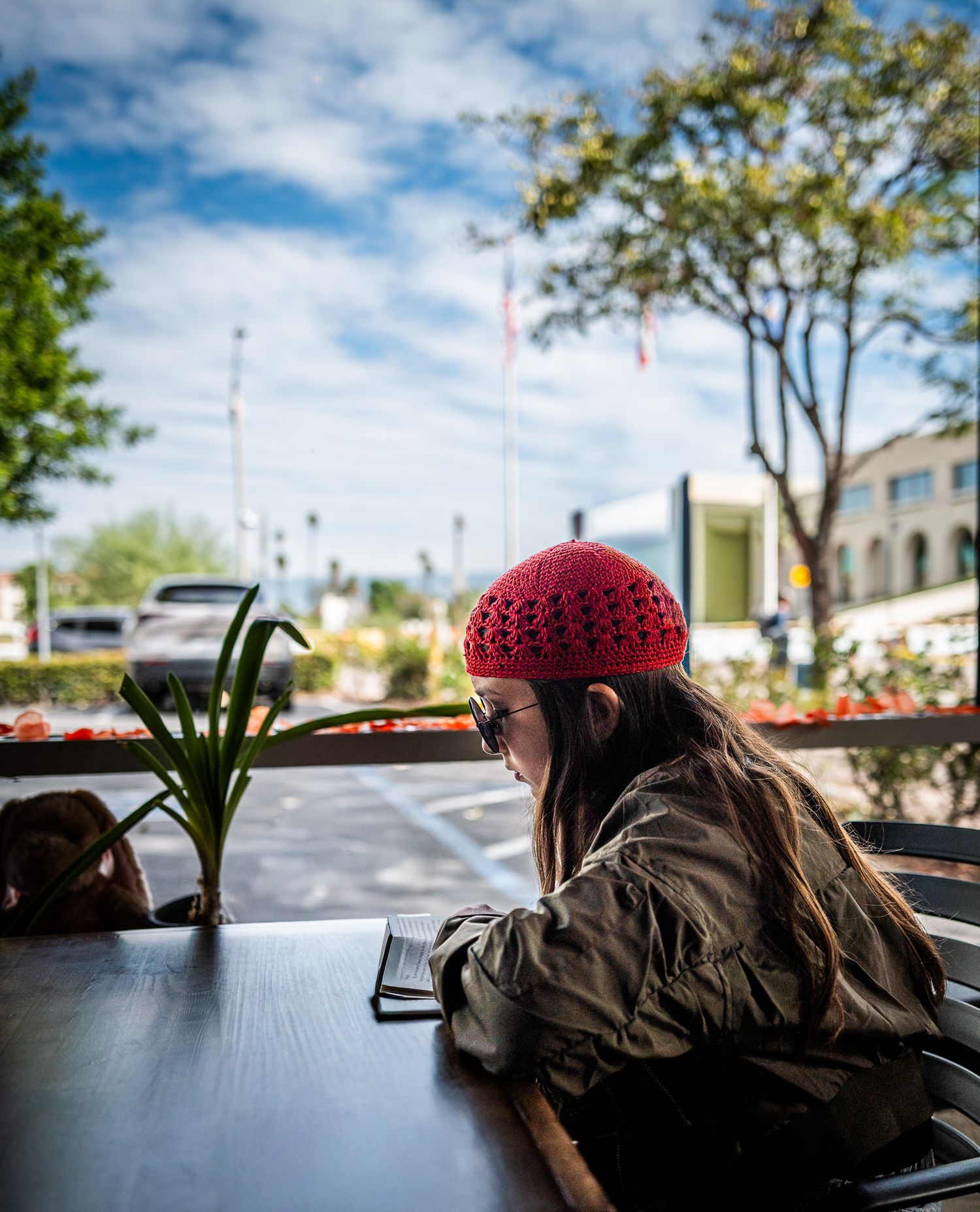 Woman in a red crocheted beanie and sunglasses sits at an outdoor cafe table, looking toward a sunny parking lot and street.
