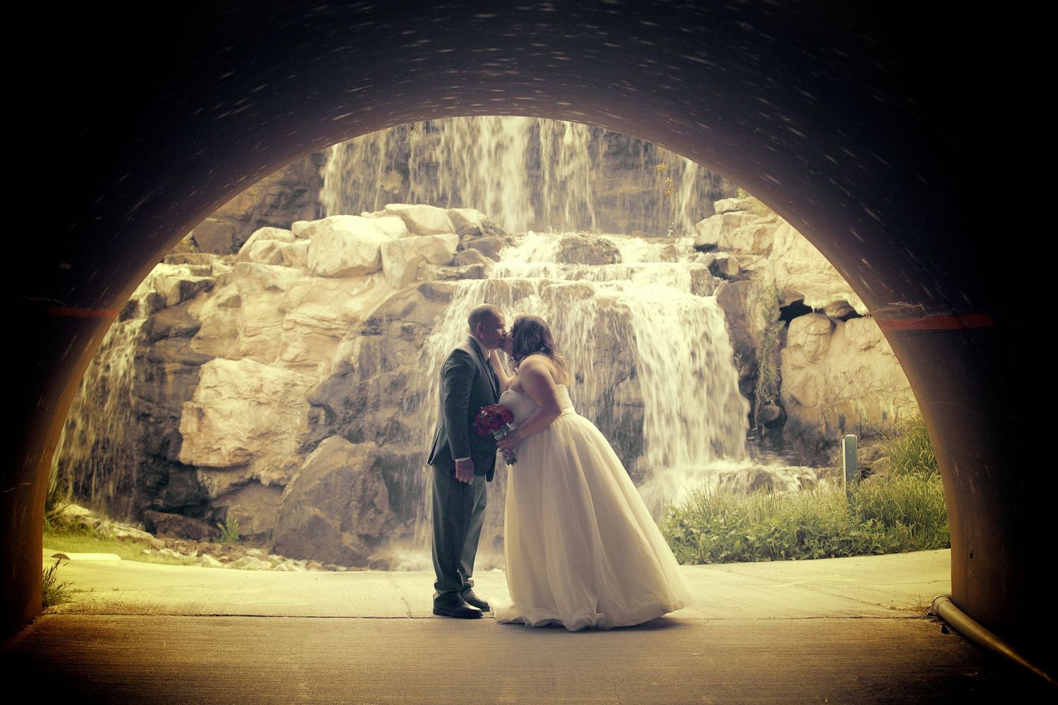 Bride and groom sharing a romantic kiss under a stone arch with a waterfall backdrop at their wedding.