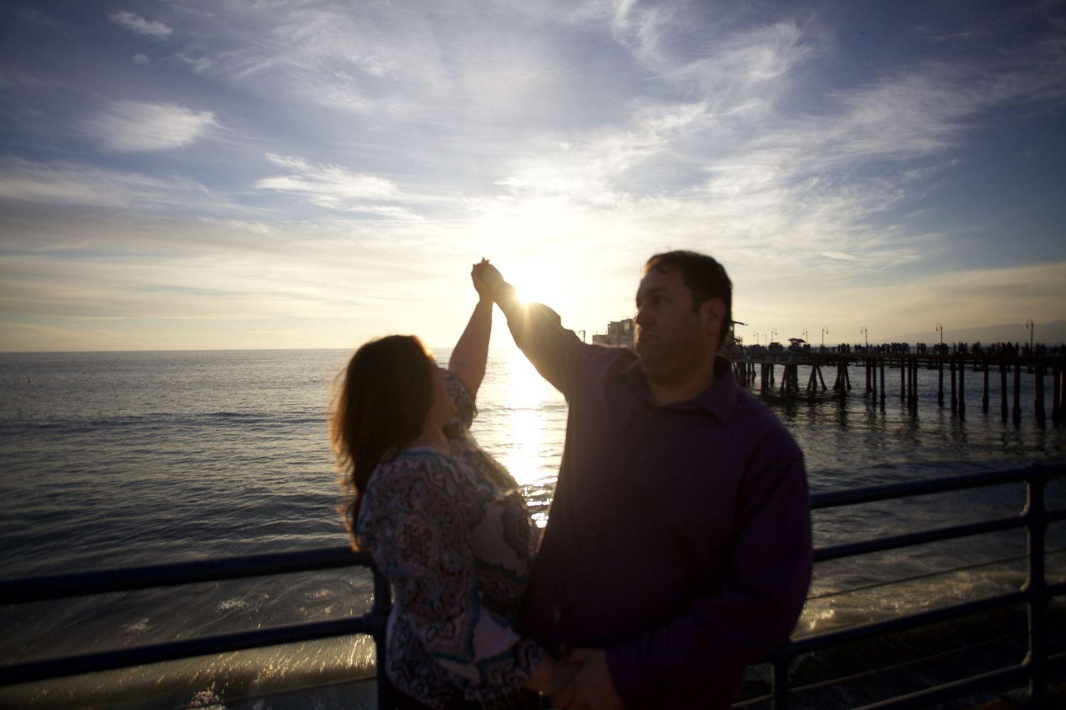A couple silhouetted against a sunset at a beach pier, holding hands raised.