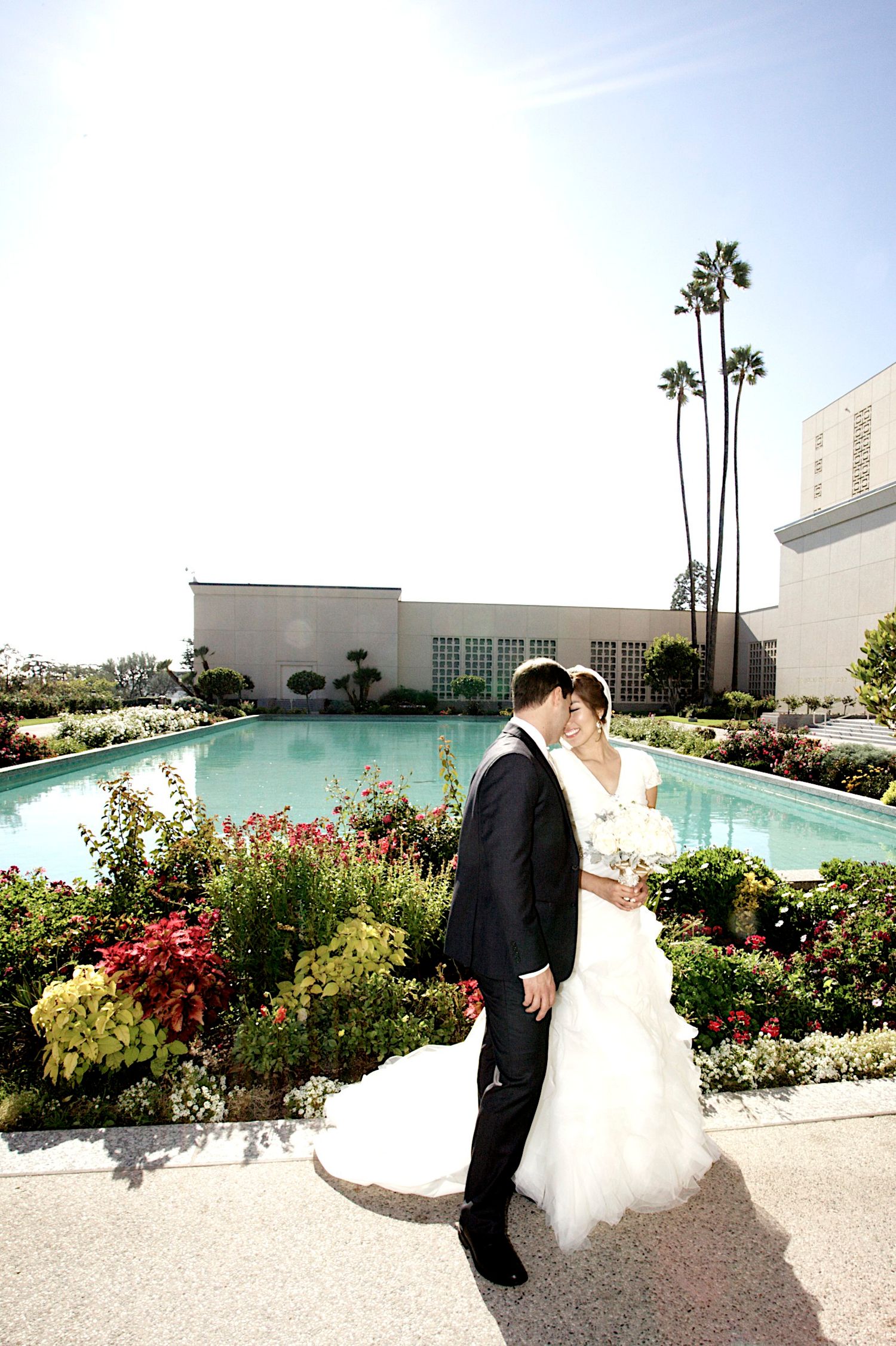 Bride and groom kissing in a lush garden with a reflecting pool and palm trees on their wedding day.