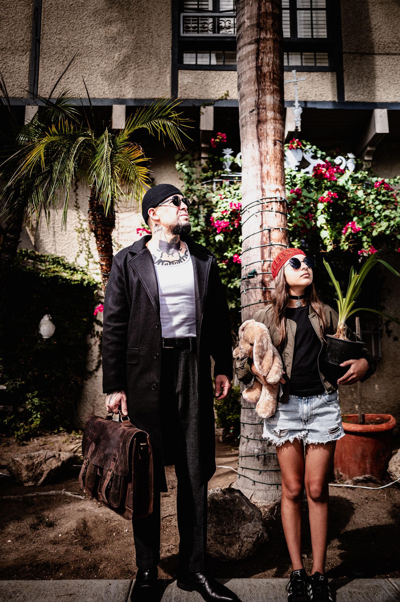 Man in a long black coat and sunglasses holding a leather briefcase beside a girl in a red beanie carrying a teddy bear under a palm tree.