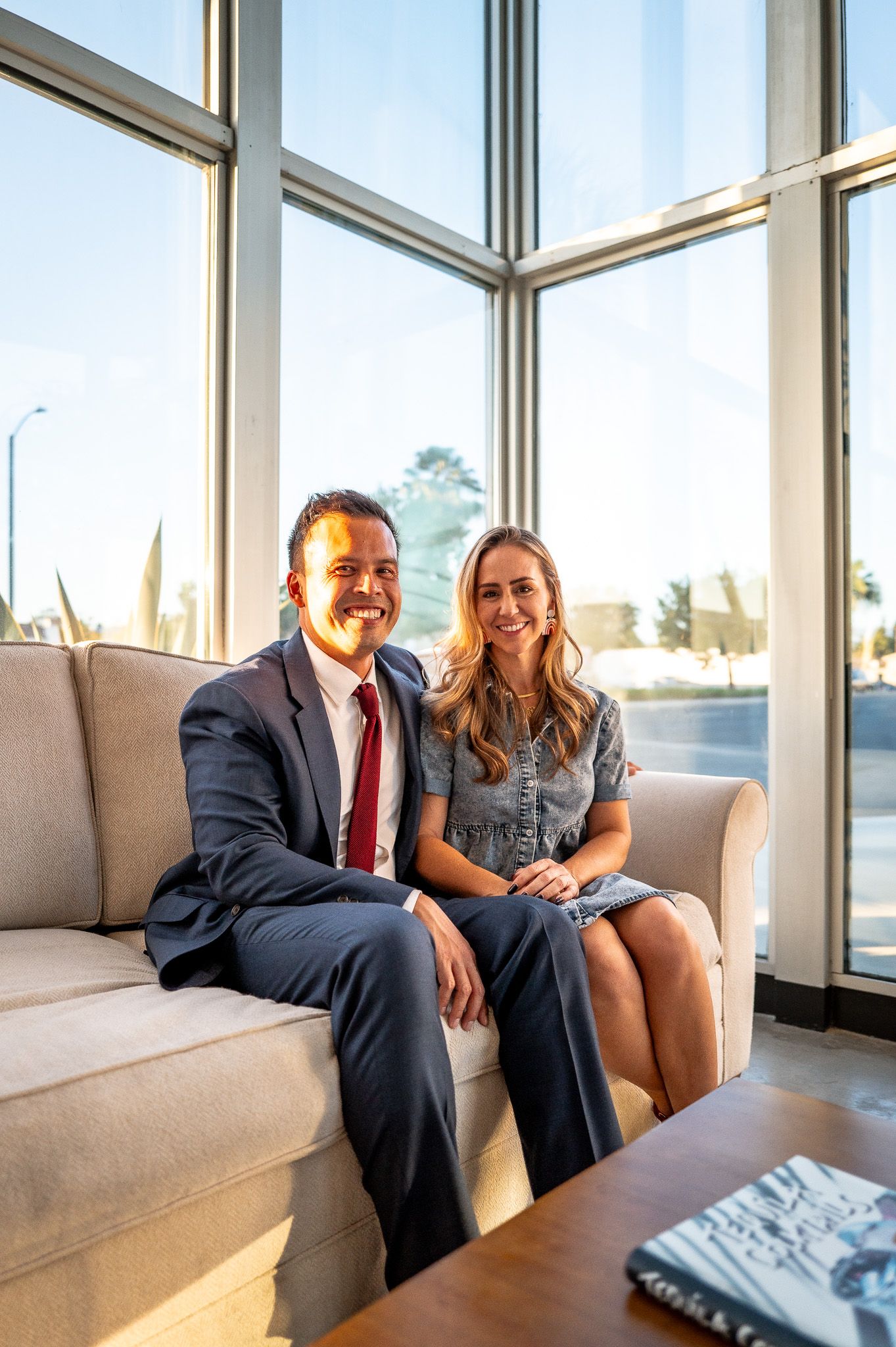 Smiling man in a navy suit and red tie sits beside a woman in a denim dress on a cream sofa in a bright, window-lined room.
