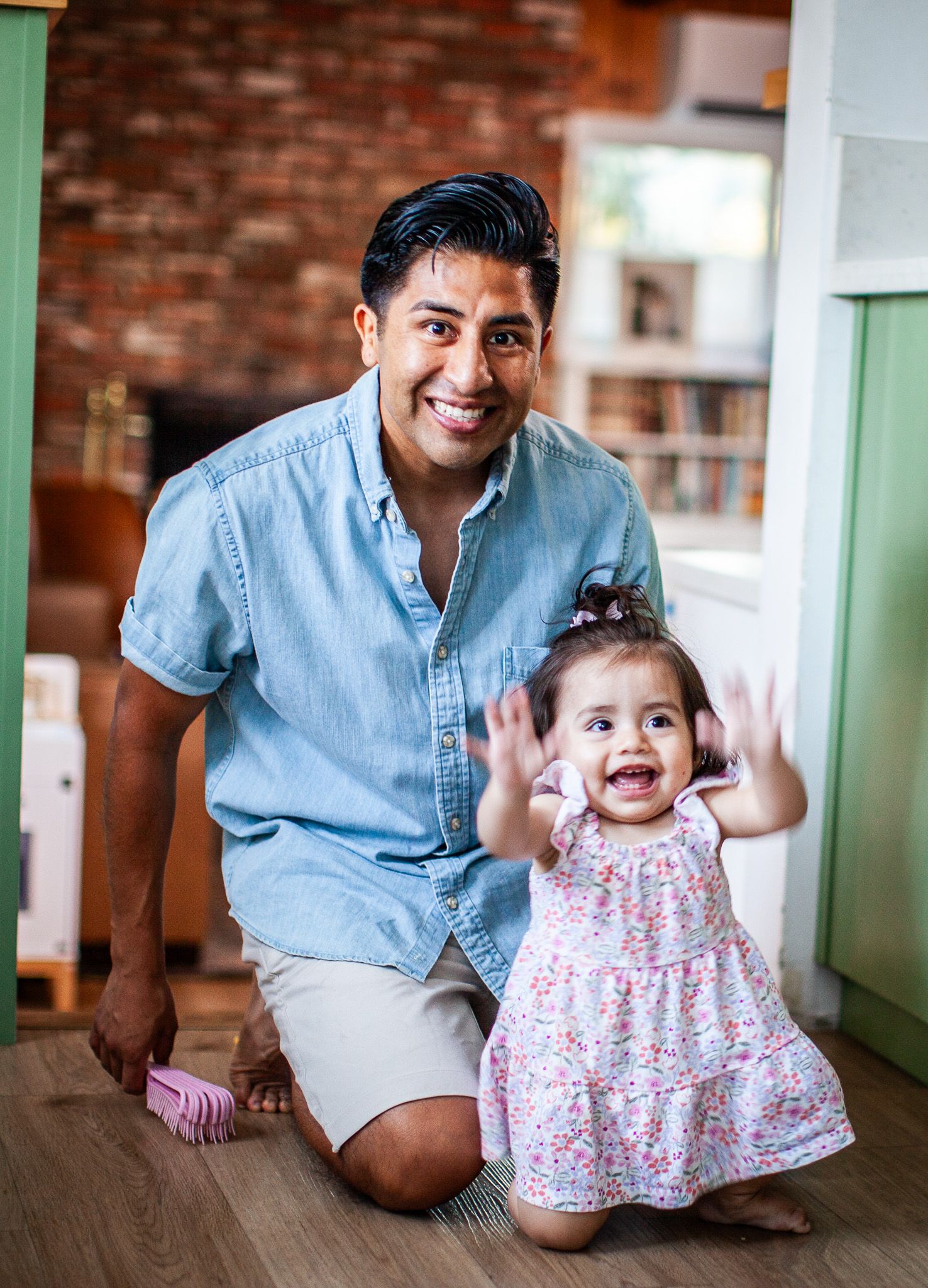A smiling man kneels on a wooden floor beside a happy toddler girl in a floral dress, both posing indoors.